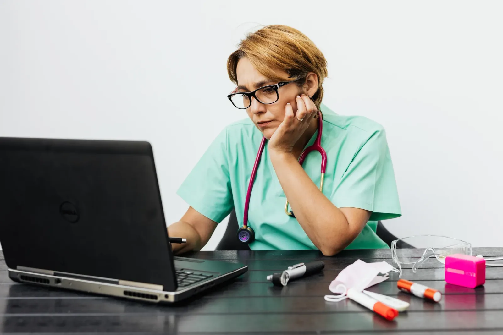 A nurse wearing eyeglasses works at a laptop on a desk, with medical instruments beside the keyboard.